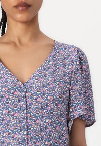 Blus med blomstermotiv i blått, med korta ärmar, V-ringning och knapppynt. Mjukt tyg med varierande färger inklusive rosa och grönt.