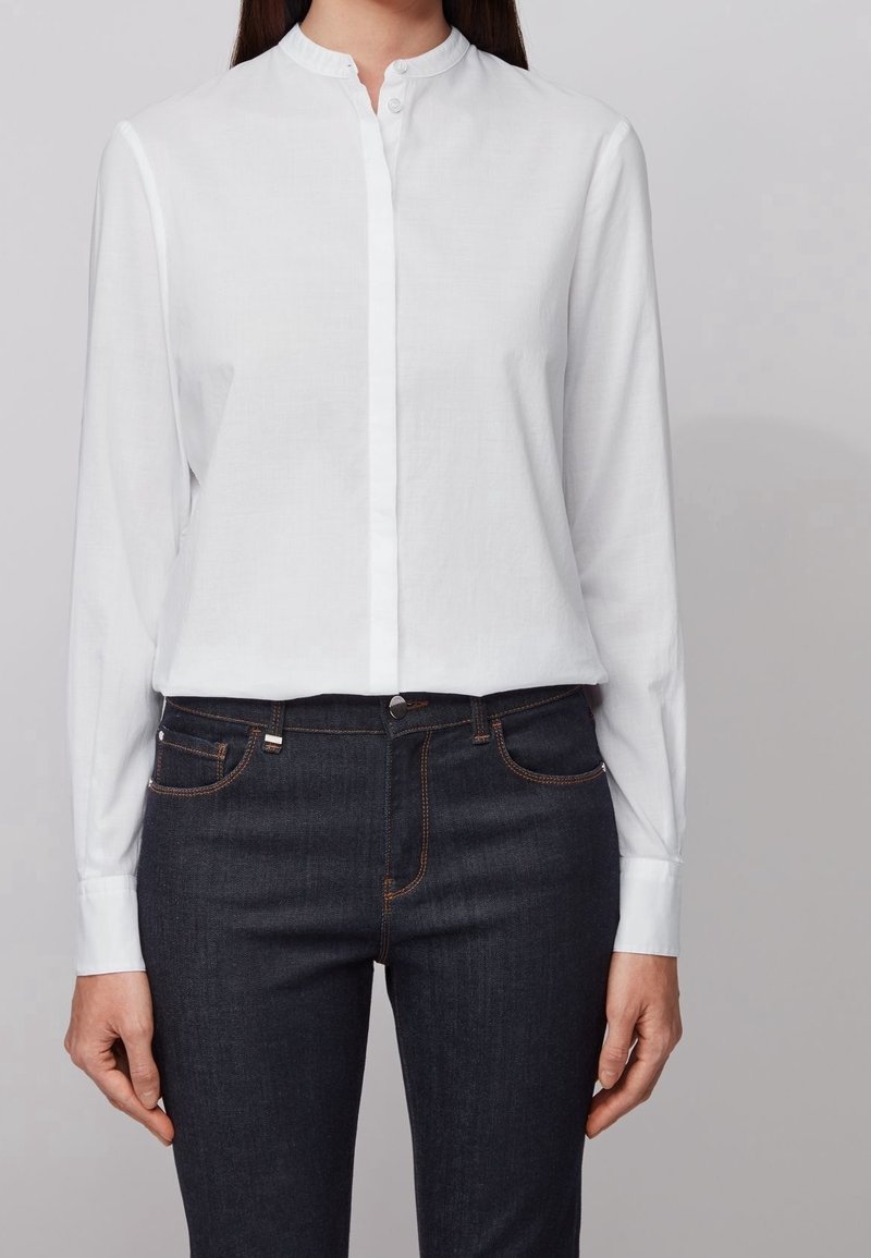 Femme portant une chemise blanche à manches longues boutonnée avec un col officier et un jean bleu foncé, debout, les bras relâchés le long du corps.