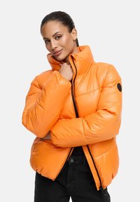 Orange puffjacka med hög krage, quiltat mönster och dragkedjestängning. Har en svart logotyp patch på vänster ärm. Strukturerad yta.