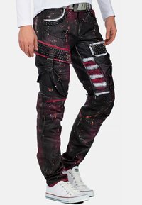 Zwarte jeans met rode en witte spatten, voorzien van studs, cargozakken en gestreepte details. Gecombineerd met witte sneakers.