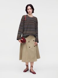 Femme portant un haut rayé à manches longues, une jupe midi beige avec des boutons, des ballerines rouges à lacets, tenant un petit sac rouge avec une bandoulière chaîne.