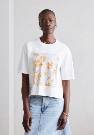 Γυναίκα με πλεξούδες που φορά λευκό μπλουζάκι με κίτρινο λουλουδάτο σχέδιο και ανοιχτό μπλε τζιν φούστα σε απλό φωτεινό φόντο.