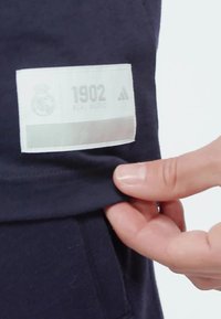 Sudadera azul marino con una etiqueta blanca que lleva el texto "1902 REAL MADRID" y el logo de Adidas, mostrando un acabado textil suave.