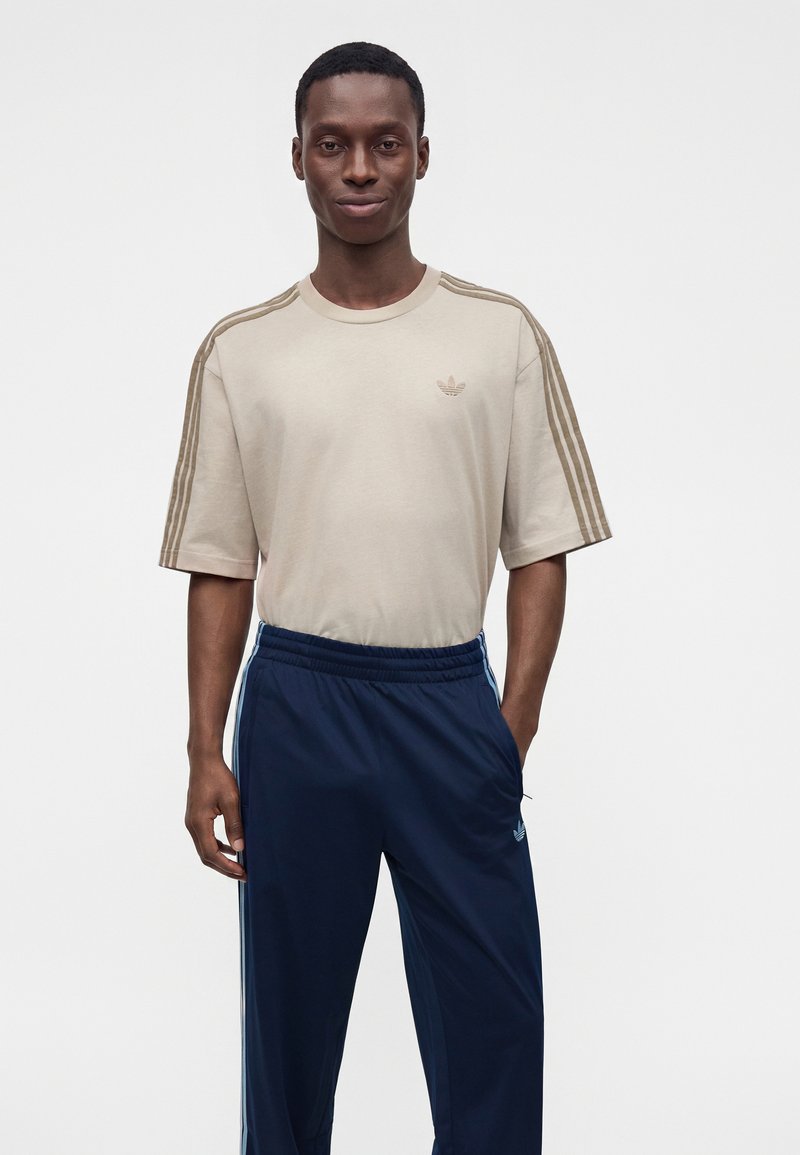 Man wearing beige Adidas T-shirt with three stripes on sleeves and navy blue Adidas track pants with light blue stripes.