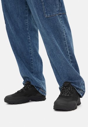 Czarne sportowe trampki z teksturowaną tkaniną i gumową podeszwą, w połączeniu z luźnymi niebieskimi dżinsami z boczną kieszenią.