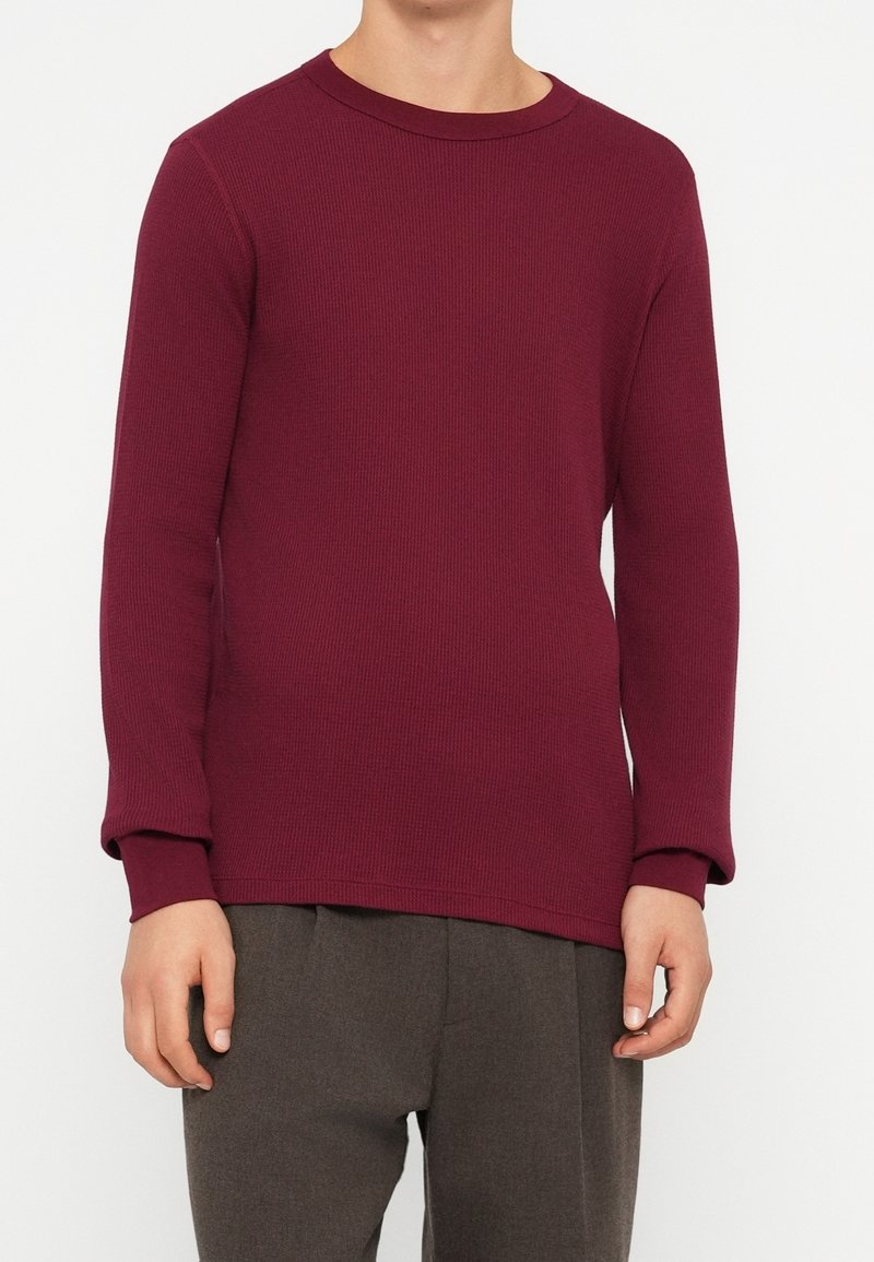 Personne portant un pull bordeaux côtelé à manches longues associé à un pantalon gris foncé, debout devant un fond uni.