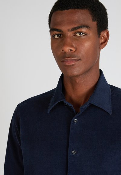 Jonge man met kort krullend haar, gekleed in een donkerblauw corduroy overhemd met grote kraagknopen, tegen een eenvoudige lichte achtergrond.