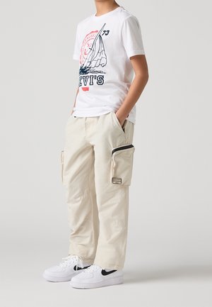 Vit t-shirt med en segelbåtsgrafik och texten "LEVI'S", tillsammans med krämfärgade cargo-byxor med sidofickor och vita sneakers.