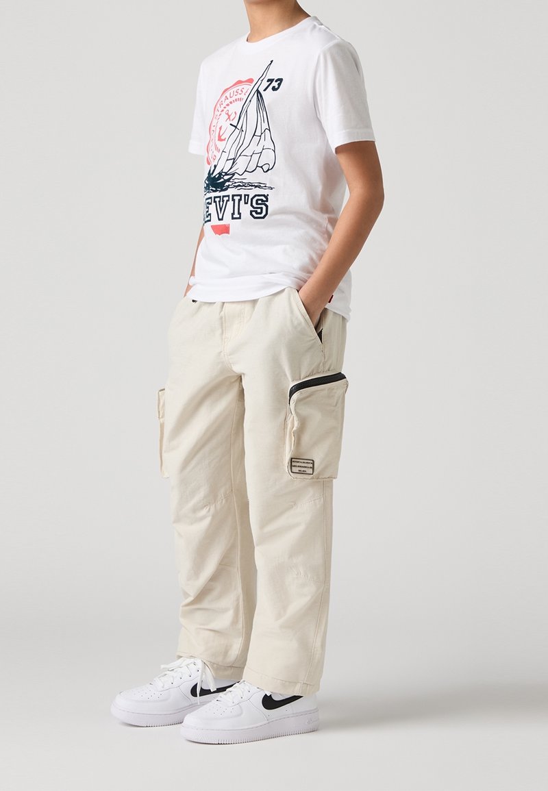 Vit t-shirt med en segelbåtsgrafik och texten "LEVI'S", tillsammans med krämfärgade cargo-byxor med sidofickor och vita sneakers.