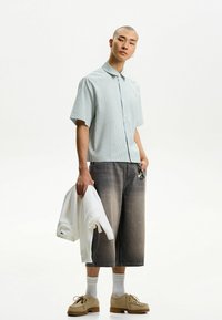 Kortærmet stribet skjorte med krave i lyseblå, parret med grå vide shorts. Modellen holder en hvid sweatshirt og har brune sko på.