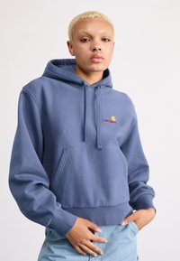 Sweat à capuche bleu clair, en tissu doux, avec une poche avant et un logo brodé en rouge et jaune.