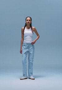 Canotta bianca, jeans azzurri chiari e sneakers bianche. Il modello è in piedi contro uno sfondo blu pallido, mostrando un abbigliamento casual con una vestibilità rilassata.