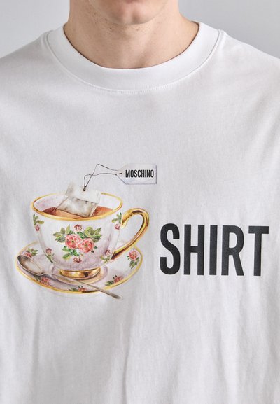 Valge särk, millel on värviline teetassi graafika lillemotiividega ja kuldsete aktsentidega, mille kõrval on suur must tekst "SHIRT."