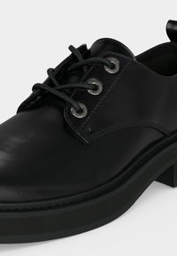 Chaussure noire en cuir à lacets avec semelle épaisse et bout arrondi, comportant trois œillets métalliques de chaque côté.