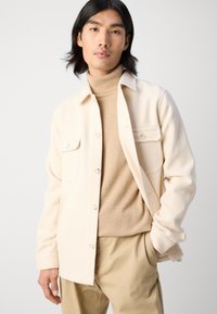 Cream, textured button-up jacket with two chest pockets, worn over a beige turtleneck. Paired with light beige trousers.