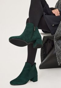 Tmavě zelené semišové kotníkové boty se zipem, mandlovou špičkou a blokovým podpatkem, umístěné na černém oblečení a kontrastním texturovaném pozadí.