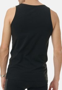 Man som bär en åtsittande svart ärmlös topp och svarta byxor, stående med ryggen mot kameran mot en vit bakgrund.