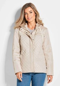 Beige gesteppte Jacke mit Kapuze, ausgestattet mit einem Reißverschluss vorne, Druckknopfverschlüssen und zwei seitlichen Taschen. Glatte Textur und leichtes Material.