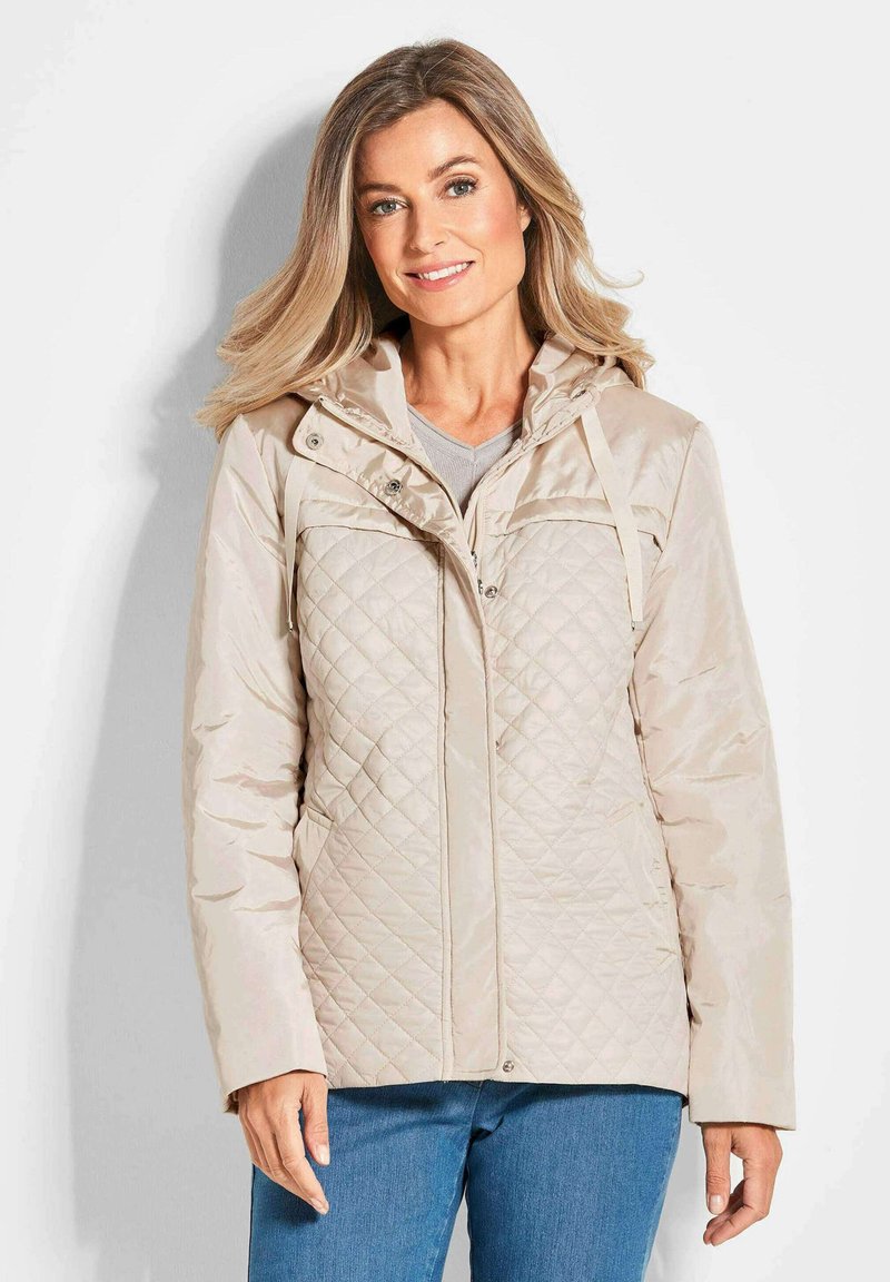 Beige gesteppte Jacke mit Kapuze, ausgestattet mit einem Reißverschluss vorne, Druckknopfverschlüssen und zwei seitlichen Taschen. Glatte Textur und leichtes Material.