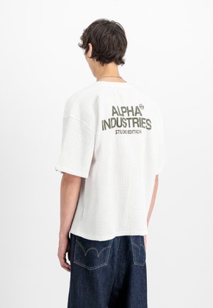 Person mit kurzen braunen Haaren trägt ein weißes, strukturiertes T-Shirt mit dem Aufdruck "ALPHA INDUSTRIES STUDIO EDITION" auf dem Rücken und dunk Blau Jeans.