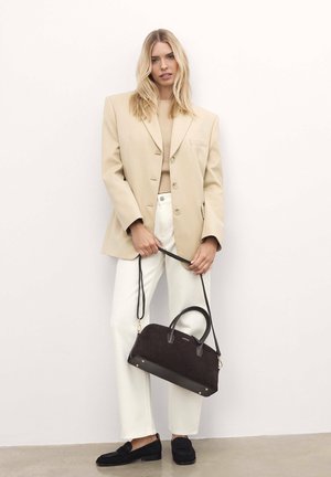 Blazer beige, maglione crema, pantaloni bianchi e mocassini neri; tiene una borsa in suede marrone scuro con dettagli in pelle e tracolla.