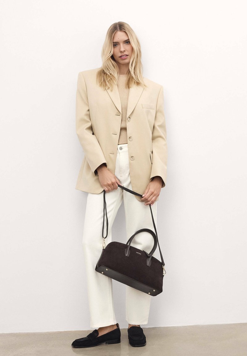 Beige blazer, cream sweater, white trousers, and black loafers; holding a dark brown suede handbag with leather accents and a shoulder strap.
