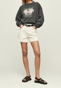 Sudadera gris con estampado gráfico, pantalones cortos de mezclilla crema con dobladillo deshilachado y sandalias negras con tiras. Cinturón negro acentúa el atuendo.