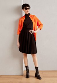 Abito nero midi con gonna svasata, abbinato a una camicia button-up arancione. Stivaletti neri con tacco; occhiali da sole neri e orecchini a cerchio.