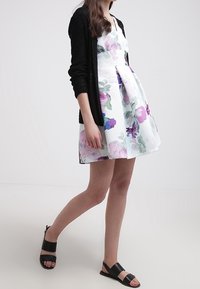 Robe florale blanche avec des accents violets et verts, tissu texturé, jupe évasée, associée à un gilet noir et des sandales plates noires.