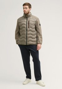 Padded jacket in olive green with a quilted design, high collar, and zippered pockets. Paired with dark pants and white sneakers.