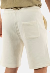 Pantaloni corti in cotone beige con vita elasticizzata, tasche laterali e una singola tasca posteriore a toppa. Morbida texture e lunghezza fino a metà coscia.