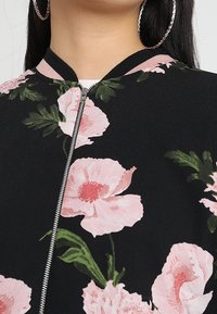 Giacca bomber floreale nera con grandi stampe di peonia rosa. Presenta una chiusura con zip e un colletto e polsini a coste neri. Texture morbida.