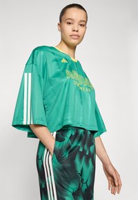 Green cropped sports top with yellow accents and Adidas logo; features white stripes on sleeves. Paired with patterned green and black pants.