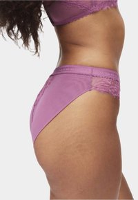 Kobieta w fioletowej koronkowej bieliźnie, pokazana od boku od talii do górnej części uda, podkreślająca wzór i dopasowanie na jasnym tle.