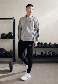 Siva zip-up bunda z črnimi črtami, črne hlače in bele čevlje. Na bundi in hlačah je logotip Adidas. V ozadju so fitnes naprave.