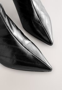 Bottes en cuir noires brillantes à bout pointu avec un motif texturé subtil, montrées en gros plan sur un fond clair.
