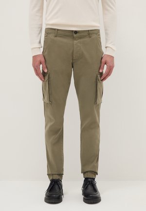 Uomo che indossa pantaloni cargo verde oliva con polsini elastici, una camicia a maniche lunghe color crema e scarpe nere con lacci, in piedi davanti a uno sfondo semplice.