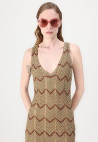 Abito in maglia con scollatura a V profonda, caratterizzato da un motivo a zigzag in oro e borgogna. Il modello indossa occhiali da sole oversize rosa, con le mani nelle tasche.