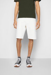 Shorts en coton blancs avec une coupe droite, associés à un t-shirt vert foncé. Baskets montantes noires et blanches avec des semelles en caoutchouc gomme.