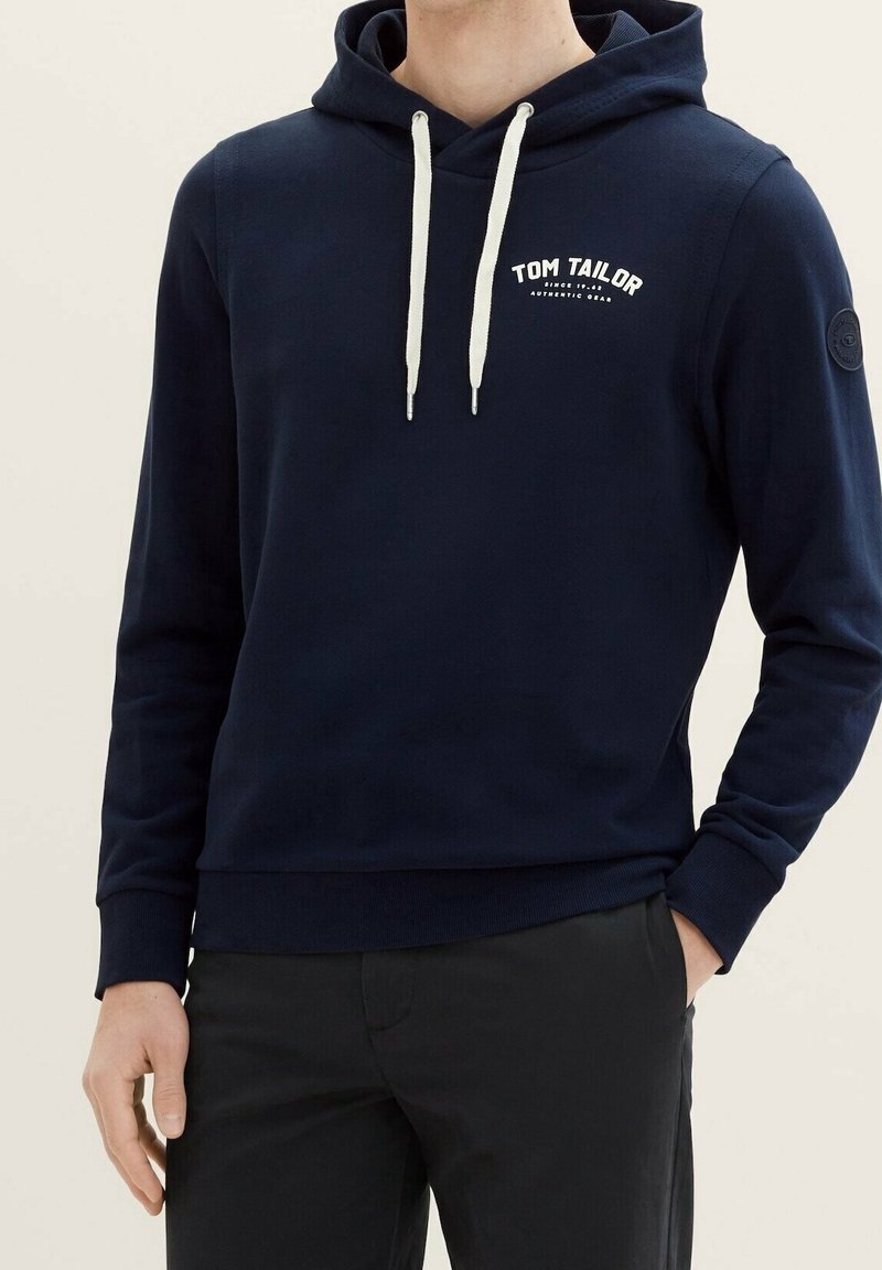Granatowa bluza z kapturem z miękkiego materiału z białymi sznurkami. Posiada nadrukowane logo na klatce piersiowej oraz okrągłą naszywkę z logo na rękawie.
