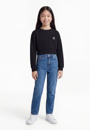 Fille souriante aux longs cheveux noirs portant un sweat-shirt court noir Adidas, un jean taille haute bleu et des baskets blanches, debout devant un fond blanc.