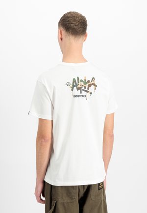 Man wearing white t-shirt with camo graffiti-style "Alpha Industries" logo on back, paired with olive cargo pants, standing against white background.