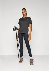 Conjunto desportivo composto por uma t-shirt preta e leggings azuis com detalhes em painéis. A modelo segura um bastão de trekking, calçando sapatos de caminhada cinzentos.