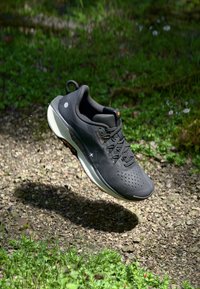 Chaussure de sport noire avec une tige en maille textur�ée, semelle rembourrée, design à lacets et accents de logo subtils, flottant au-dessus d'une surface herbeuse.