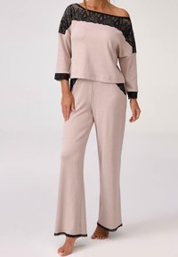 Jasno beżowy zestaw odzieży do wypoczynku z czarnymi koronkowymi akcentami. Posiada top na jedno ramie oraz szerokie spodnie. Miękka tekstura, luźny krój i starannie wykończone krawędzie.