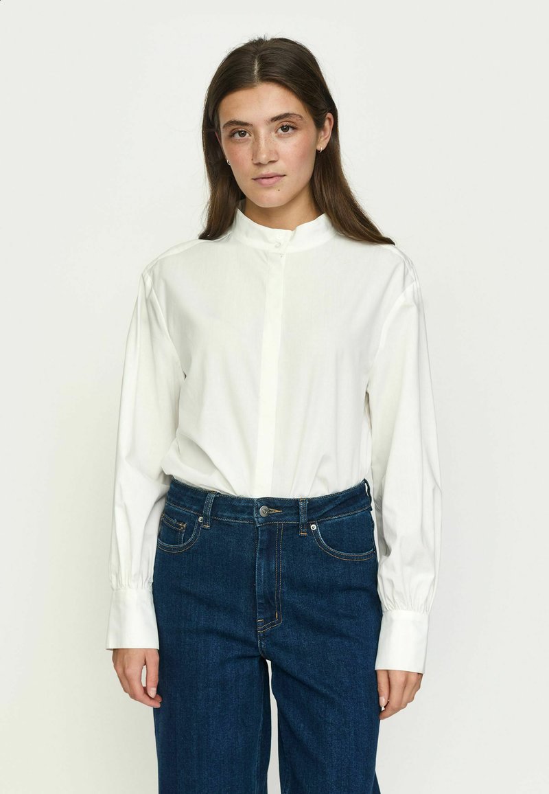 Femme aux longs cheveux bruns portant une chemise blanche à manches longues boutonnée et un jean bleu, debout devant un fond uni clair.