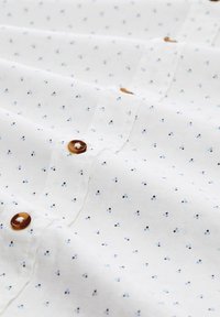 Chemise en coton blanche avec de petits pois multicolores. Boutons marron à finition lisse sur le devant. Texture douce et design classique.