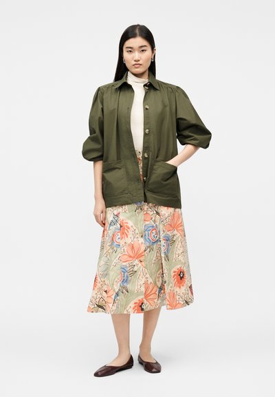 Γυναίκα με κοτλέ μπουφάν σε ελιά πράσινο χρώμα πάνω από μπεζ ζιβάγκο και πολύχρωμη φλοράλ μίντι φούστα, φορεμένη με καφέ φλατ παπούτσια, στέκεται μπροστά σε λευκό φόντο.