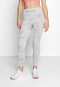 Šedé maskáčové legíny s vysokým pasem, na kterých je logo Puma. Materiál je elastický a přiléhavý, v kombinaci s bílými sportovními botami.