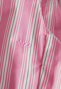 Růžová tkanina s bílými a černými vertikálními pruhy. Obsahuje vyšitý nápis "JUXX" růžovou nití. Textura se zdá být hladká a lehká.